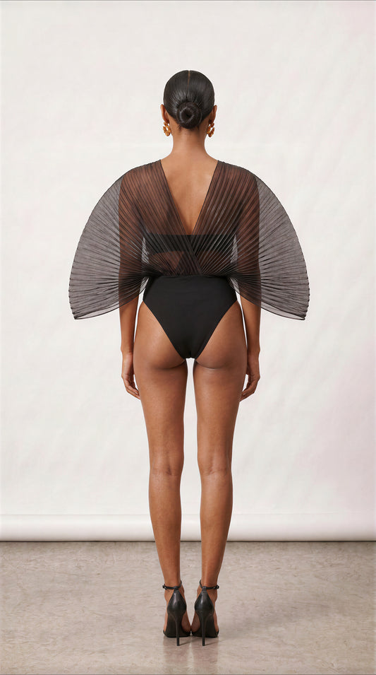 A model shown from the back wearing a black one-piece swimsuit with a sheer, pleated cape-like back overlay and a deep V-back, paired with black high heels against a neutral studio backdrop.