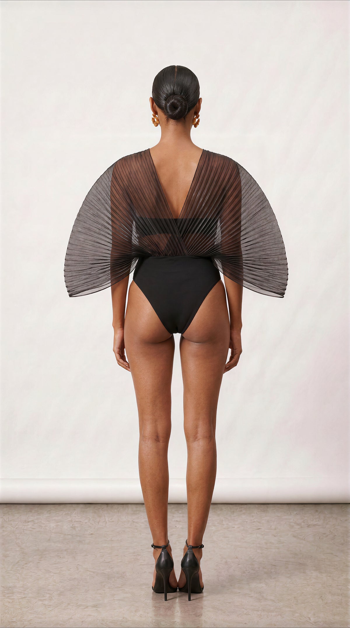 A model shown from the back wearing a black one-piece swimsuit with a sheer, pleated cape-like back overlay and a deep V-back, paired with black high heels against a neutral studio backdrop.