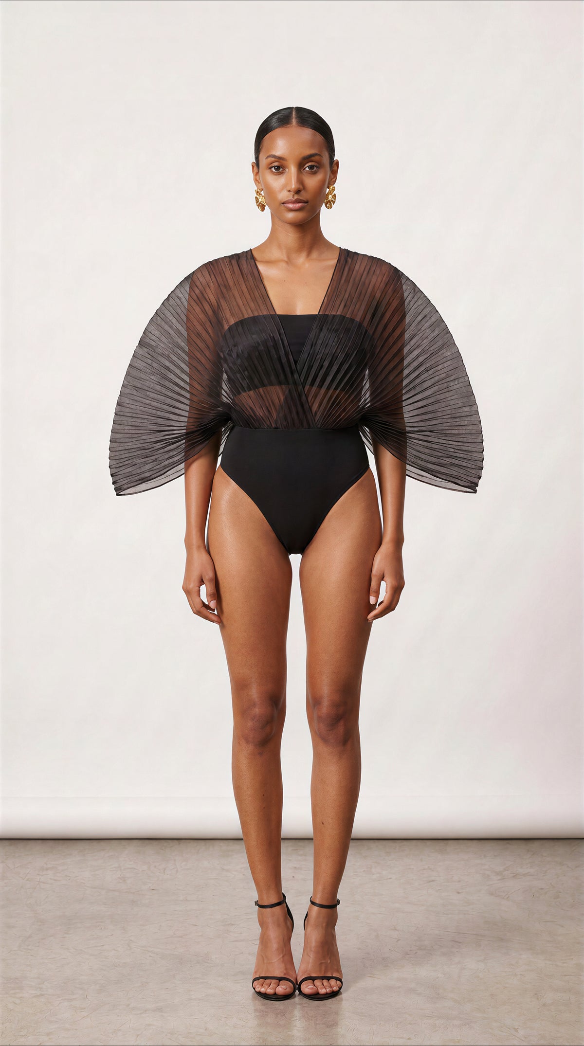 A model stands against a plain light studio background wearing a black one-piece swimsuit with dramatic sheer pleated cape sleeves and a square neckline.
