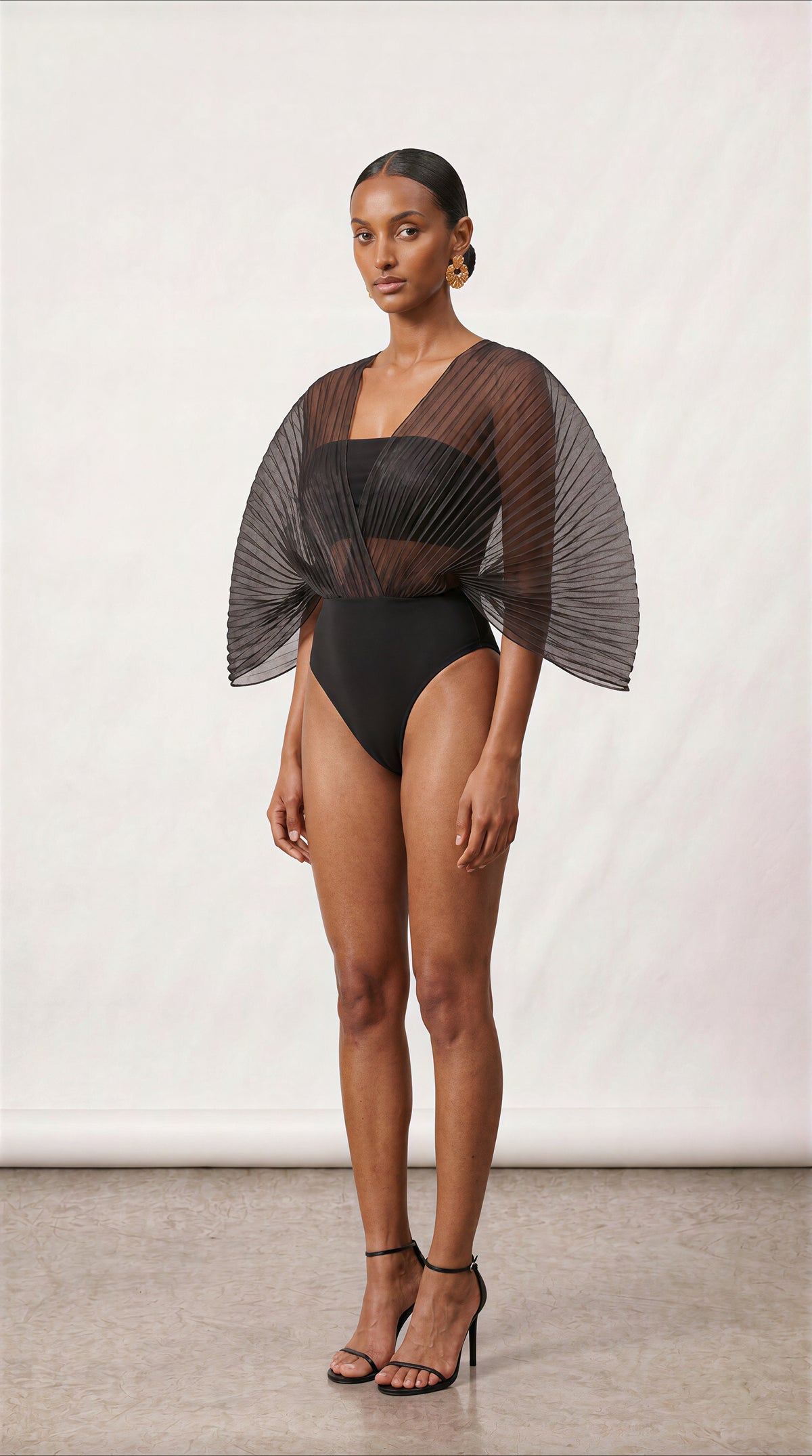 A model stands against a light neutral studio backdrop wearing a black one-piece swimsuit with dramatic sheer pleated cape sleeves and a square neckline.