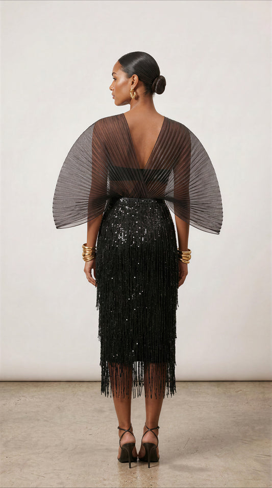 A model is shown from the back wearing a black pleated fringe midi skirt with a sheer black pleated cape-style top overlay, set against a neutral studio background.